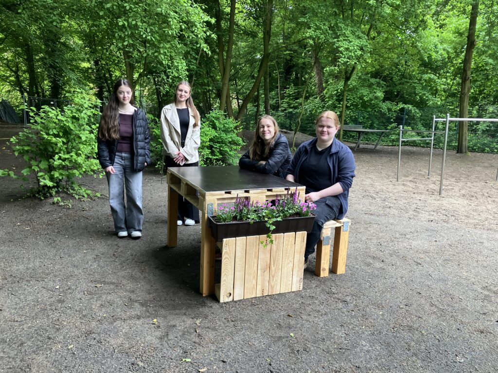 Eine Gruppe von Mädchen sitzt auf einer selbst gezimmerten Bank-Tisch-Kombination, an den Enden befinden sich Beetelemente