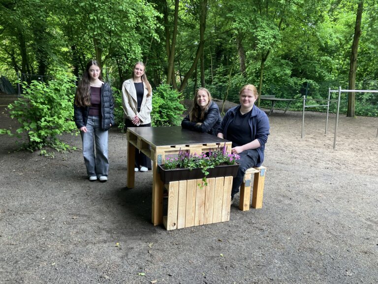 Eine Gruppe von Mädchen sitzt auf einer selbst gezimmerten Bank-Tisch-Kombination, an den Enden befinden sich Beetelemente