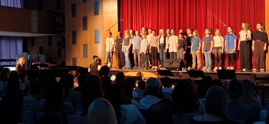 Der kleine Chor steht beim kleinen Sommerkonzert auf der Bühne und singt. Herr Dr. Kandt begleitet am Klavier und Frau Inselmann dirigiert.