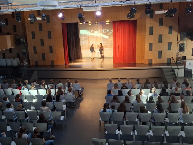 Theateraufführung in der Aula, auf der Bühne performen drei Schülerinnen