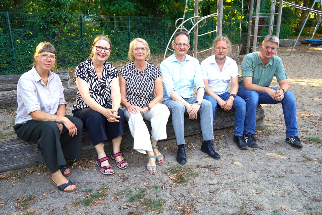 Die Schulleitungsrunde sitzt auf einem Holzstamm auf dem Dobbenhof und lächelt freundlich in die Kamera